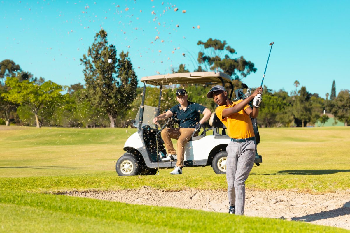 Caucasian young man sitting in cart looking at african american friend playing golf against sky. copy space, summer, transportation, unaltered, friendship, togetherness, sport, weekend activities.