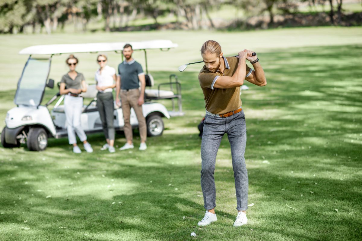 Handsome man swinging with putter, playing golf on the course with friends and golf car on the background on a sunny weather